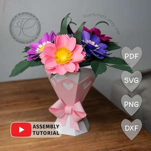 May include: A paper flower bouquet in a pink and white vase with a bow. The bouquet features pink, purple, and blue flowers with yellow centres and green leaves. The vase has heart cutouts. The image includes the text "Assembly Tutorial".