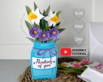 Puede incluir: Un arreglo floral de papel en forma de tarro Mason azul, con narcisos blancos con centros amarillos y flores moradas. El tarro tiene una cinta morada y el texto "Thinking of you".
