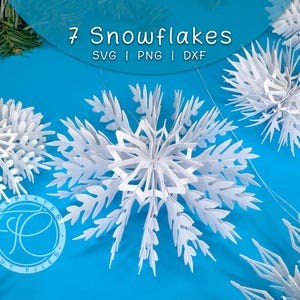 May include: White paper snowflakes with intricate designs are displayed against a bright blue background. The image includes the text "7 Snowflakes" and file type information. The snowflakes are a winter-themed craft or decoration.