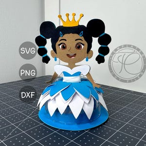 May include: A papercraft princess figure with a blue and white dress, black hair in pigtails, and a gold crown. The figure has a smiling face and blue earrings. The image also includes the text "SVG", "PNG", and "DXF".