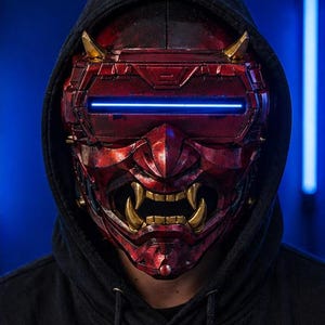 May include: A red and gold Oni mask with a blue light bar across the eyes. The mask has sharp teeth and horns, and is worn with a black hooded sweatshirt. The mask is a cosplay or costume item.