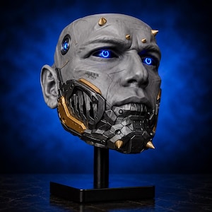 May include: A gray, futuristic head sculpture with glowing blue eyes and gold accents. The sculpture has a detailed, mechanical face mask and is mounted on a black stand. The head has a menacing expression and is set against a dark blue background.