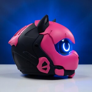 May include: A futuristic helmet in black and bright pink, shaped like a cat's head. It has a glowing blue eye and cat ears. The design is modern, with geometric shapes and a vibrant colour scheme. The helmet is likely for cosplay or display.
