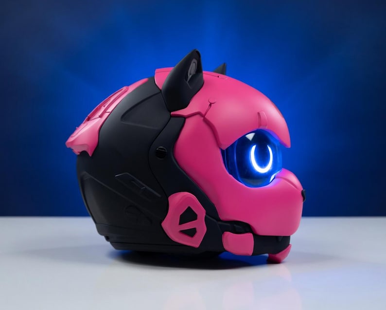 May include: A futuristic helmet in black and bright pink, shaped like a cat's head. It has a glowing blue eye and cat ears. The design is modern, with geometric shapes and a vibrant colour scheme. The helmet is likely for cosplay or display.