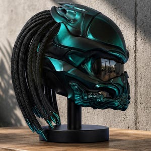 Può includere: Un casco metallico color verde acqua e nero caratterizzato da un design organico scolpito con una superficie liscia e sagomata. Il casco include lunghi cordini neri intrecciati che si estendono dal retro e una visiera scura riflettente.