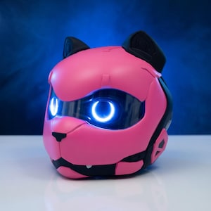 May include: A pink robotic cat head with black detailing and ears. The cat's eyes emit a bright blue light. The design has a futuristic aesthetic, with smooth, rounded surfaces and a sleek appearance. The background is a gradient of blue.