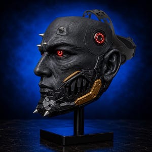 Op de afbeelding: Een gedetailleerd zwart masker met rood oplichtende ogen en gouden metalen accenten. Het masker heeft scherpe spikes en een complex ontwerp, gemonteerd op een zwarte standaard tegen een blauwe achtergrond. Het masker heeft een dreigende uitstraling.