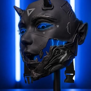 Op de afbeelding: Een zwart, futuristisch masker met gloeiende blauwe accenten, dat lijkt op een gestileerd gezicht met hoorns. Het masker heeft ingewikkelde details en is gemonteerd op een zwarte standaard, tegen een blauwe achtergrond.