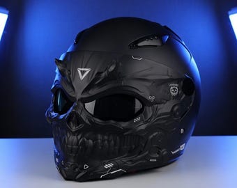 May include: A matte black motorcycle helmet with a skull design. The helmet features a detailed skull face with eye sockets, a jaw, and horns. The visor is dark, and the helmet has a futuristic, edgy aesthetic.