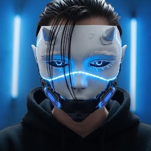 Op de afbeelding: Een futuristisch wit masker met gloeiende blauwe accenten, waaronder de ogen en een gezichtsrooster. Het masker heeft kleine hoorns en puntige oren, tegen een blauwe achtergrond. De persoon draagt een zwarte hoodie.