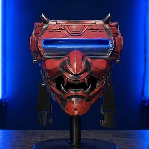 May include: A red and black futuristic mask with a menacing design, featuring a glowing blue visor. The mask is mounted on a black stand, showcasing its intricate details and cyberpunk aesthetic.