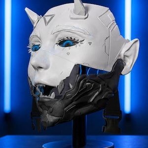 Op de afbeelding: Een wit en zwart masker met een futuristisch ontwerp, met hoorns, blauwe ogen en een gedetailleerd ondergezicht. Het masker wordt getoond op een zwarte standaard tegen een blauwe achtergrond, wat een mix van fantasie en technologie laat zien.