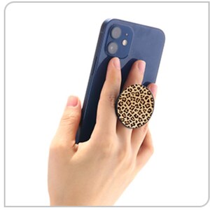 May include: A navy blue smartphone held in a hand, with a round leopard print phone grip attached. The grip features a tan and brown spotted pattern, providing a stylish and functional accessory.