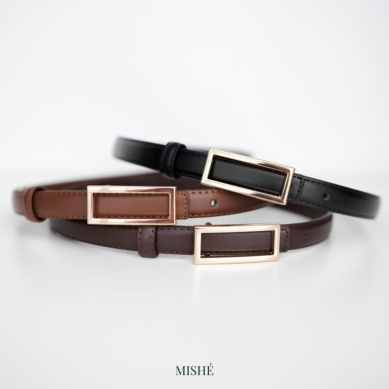 May include: Three leather belts in black, brown, and dark brown, each with a rectangular gold-colored buckle. The belts are arranged in a layered composition against a white background. The word "MISH&Eacute;" is printed at the bottom.