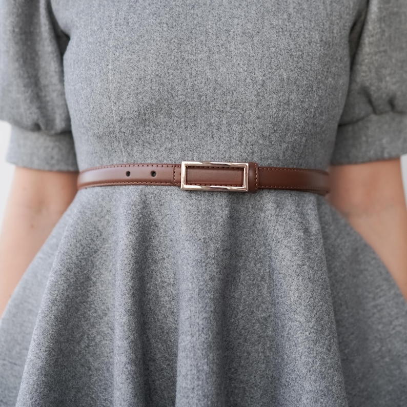 Op de afbeelding: Een bruine leren riem met een rechthoekige goudkleurige gesp wordt gedragen over een grijze jurk. De riem is dun en heeft een eenvoudig, klassiek ontwerp, waardoor de outfit een vleugje elegantie krijgt.
