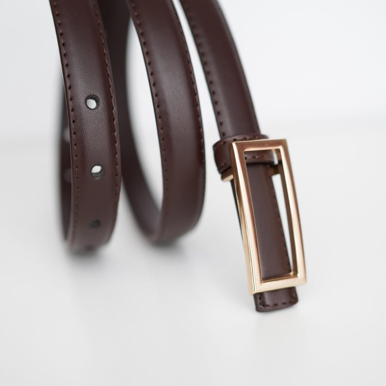 Op de afbeelding: Een bruine leren riem met een rechthoekige goudkleurige gesp. De riem is opgerold en toont de gaten voor de gesp. Het is een neutraal accessoire.