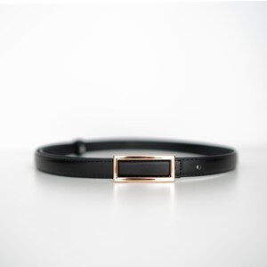May include: A black belt with a rectangular gold-colored buckle. The belt is made of a smooth, shiny material and is designed to be worn around the waist. The buckle is a simple, elegant design.