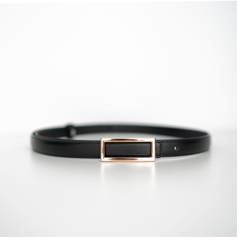 May include: A black belt with a rectangular gold-colored buckle. The belt is made of a smooth, shiny material and is designed to be worn around the waist. The buckle is a simple, elegant design.