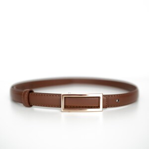 Op de afbeelding: Een bruine leren riem met een rechthoekige goudkleurige gesp. De riem is een dunne stijl, geschikt voor diverse outfits. De gesp heeft een eenvoudig, elegant ontwerp.