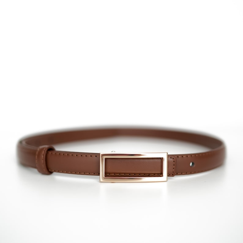 Op de afbeelding: Een bruine leren riem met een rechthoekige goudkleurige gesp. De riem is een dunne stijl, geschikt voor diverse outfits. De gesp heeft een eenvoudig, elegant ontwerp.