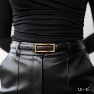 May include: A black leather belt with a gold rectangular buckle. The belt is worn with black leather pants and a black long-sleeved top. The word "MISH&Eacute;" is visible in the bottom right corner.