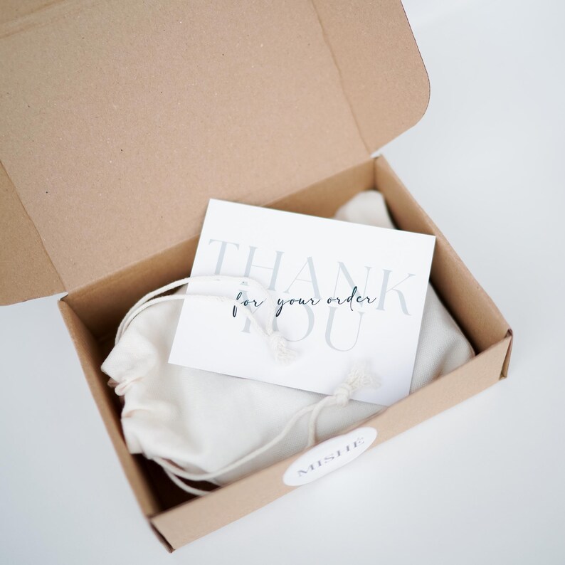 May include: An open cardboard box containing a white drawstring bag and a card that reads "THANK YOU for your order" in gray and black text. A small white sticker with the word "MISHE" is on the box.