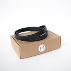 May include: A black leather belt, coiled on top of a brown cardboard box. The belt has several small holes. A white sticker with a handwritten "m" is on the box. The background is white.