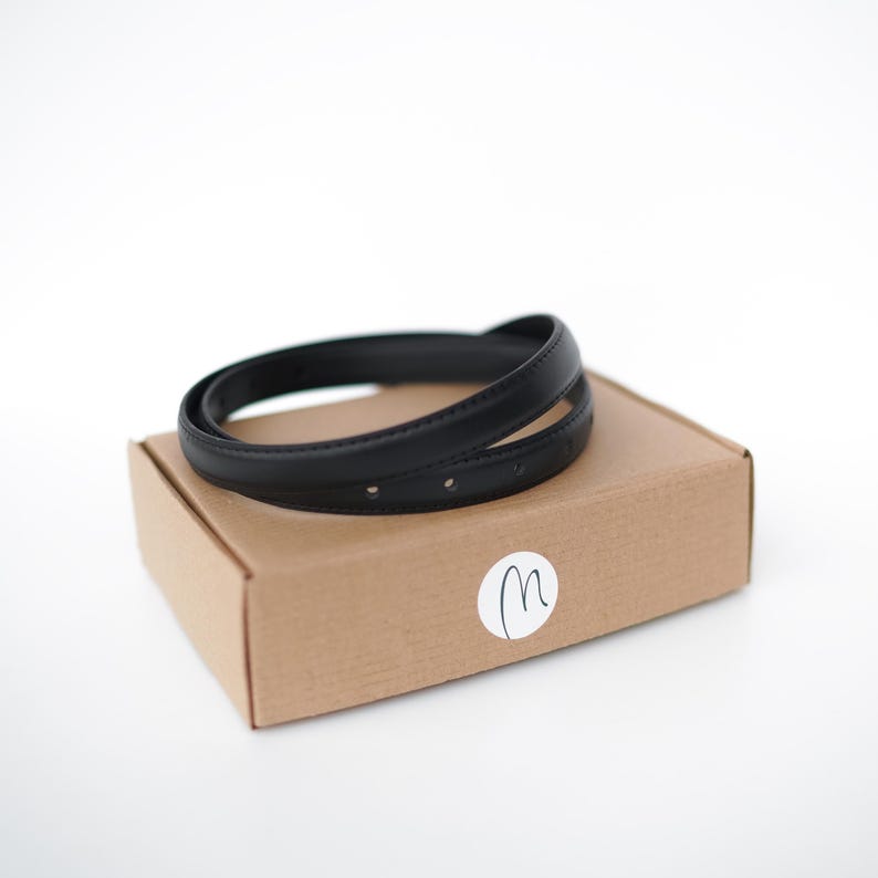 May include: A black leather belt, coiled on top of a brown cardboard box. The belt has several small holes. A white sticker with a handwritten "m" is on the box. The background is white.