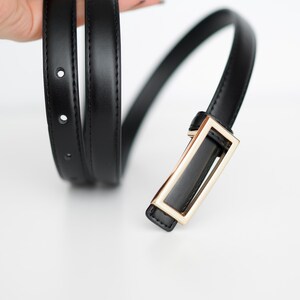 Op de afbeelding: Een zwarte riem met een rechthoekige goudkleurige gesp. De riem is gemaakt van een glad, glanzend materiaal en heeft verschillende gaten voor verstelling. De gesp heeft een eenvoudig, modern ontwerp.