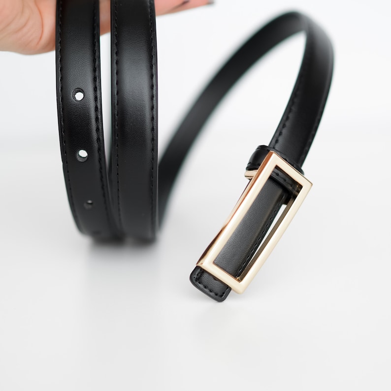 Op de afbeelding: Een zwarte riem met een rechthoekige goudkleurige gesp. De riem is gemaakt van een glad, glanzend materiaal en heeft verschillende gaten voor verstelling. De gesp heeft een eenvoudig, modern ontwerp.