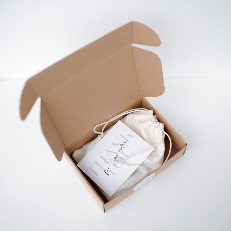 Op de afbeelding: Een open bruine kartonnen doos met een witte zak met trekkoord en een kaart met de tekst "THANK for our order". De doos staat op een witte ondergrond.