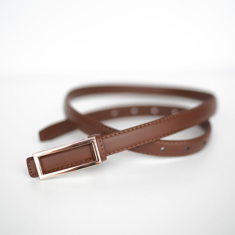 Op de afbeelding: Een bruine leren riem met een rechthoekige goudkleurige gesp. De riem is dun en heeft verschillende gaten voor verstelling. De gesp heeft een eenvoudig, modern ontwerp. De riem wordt getoond tegen een witte achtergrond.