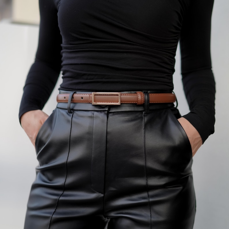 Op de afbeelding: Een bruine leren riem met een rechthoekige goudkleurige gesp. De riem wordt gedragen met een zwarte leren broek en een zwarte top met lange mouwen. De riem is ongeveer 2,5 cm breed.
