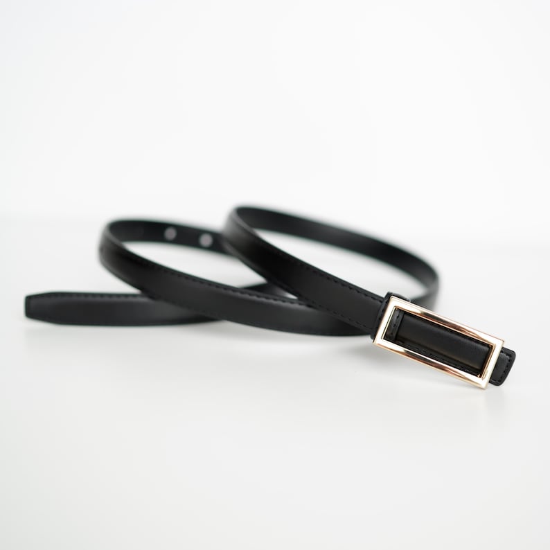 May include: A black belt with a rectangular gold-colored buckle. The belt is thin and appears to be made of leather. The belt is coiled on a white surface.