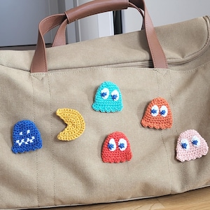 Könnte beinhalten: Eine beigefarbene Canvas-Reisetasche mit braunen Ledergriffen. Die Tasche ist mit sechs farbenfrohen, gehäkelten Pac-Man-Applikationen verziert: blaue, gelbe, orange, rote und rosa Geister sowie ein gelber Pac-Man.