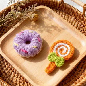 Könnte beinhalten: Nahaufnahme von zwei gehäkelten Lebensmitteln auf einem Holztablett. Eines ist ein lila Donut mit rosa und gelben Streuseln. Das andere ist ein orangefarbener Lutscher mit einer weißen Spirale und grüner Schleife.