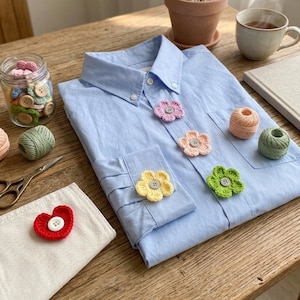 May include: A light blue button-down shirt decorated with several small, multi-colored crocheted flower appliques, each featuring a central button. The shirt is surrounded by crafting supplies including yarn balls, a jar of buttons, and scissors.