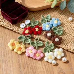 Op de afbeelding: Een verzameling kleurrijke, gehaakte bloemapplicaties in verschillende kleuren, waaronder rood, blauw, geel, roze, bruin en wit. Elke bloem heeft een contrasterend midden en groene bladeren. De bloemen zijn gerangschikt op een houten oppervlak.