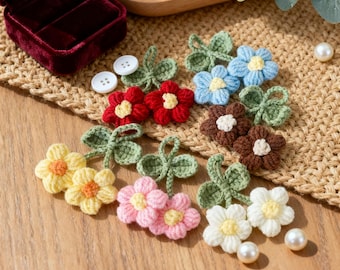 May include: A collection of colorful, crocheted flower appliques in various colors, including red, blue, yellow, pink, brown, and white. Each flower has a contrasting center and green leaves. The flowers are arranged on a wooden surface.