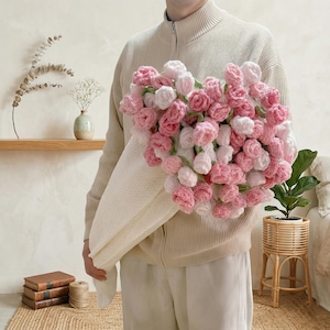 Puede incluir: Un gran ramo de flores de ganchillo en tonos rosa y blanco, envuelto en una tela color crema. Las flores hechas a mano están muy juntas, creando una apariencia llena y texturizada. El ramo lo sostiene una persona que lleva un suéter y pantalones color crema.