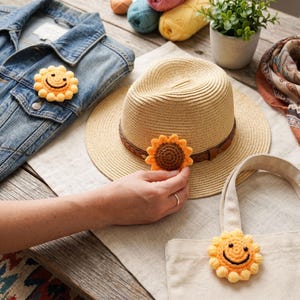 May include: A collection of handmade crochet sunflower pins and appliques. One pin is attached to a denim jacket, another to a straw hat, and a third to a canvas tote bag. The sunflowers are yellow and orange with smiling faces.