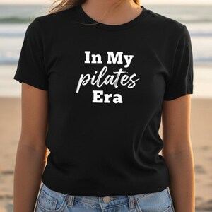 Può includere: Una t-shirt nera a maniche corte con la scritta bianca "In My pilates Era" stampata sul davanti. La maglietta presenta un girocollo e una vestibilità standard, con la parola "pilates" scritta in corsivo tra le parole "In My" ed "Era".