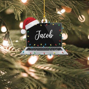 May include: A Christmas tree ornament shaped like a laptop, with the name "Jacob" on the screen. The laptop is decorated with colorful Christmas lights and a Santa hat. The ornament hangs from a gold string.