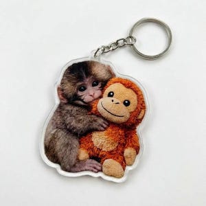 May include: A clear acrylic keychain featuring a brown and tan monkey hugging an orange plush monkey. The keychain has a silver metal ring and chain. The design is a cute and playful illustration.