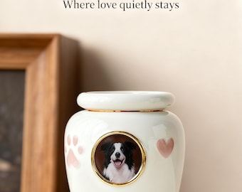 May include: A white ceramic pet urn with a gold rim and a picture of a dog. The urn has pink paw prints and hearts. The text on the urn says "Snowy Ceramic Pet Urn".