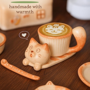 Puede incluir: Un juego de tazas de café de cerámica con temática de gato en crema y naranja, con platillo y cuchara a juego. La taza presenta un diseño de arte latte de cara de gato. El texto es "Snowy Ceramic" y "handmade with warmth".