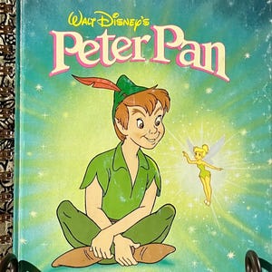 May include: A Little Golden Book titled Walt Disney's Peter Pan, featuring an illustration of Peter Pan in a green outfit with a red feather in his cap, sitting cross-legged next to a small Tinker Bell, set against a bright teal background.