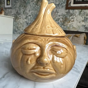 May include: A tan ceramic onion-shaped container with a lid. The container has a face with closed eyes and tear streaks. The lid is shaped like the top of an onion and has the word "Onions" on it.