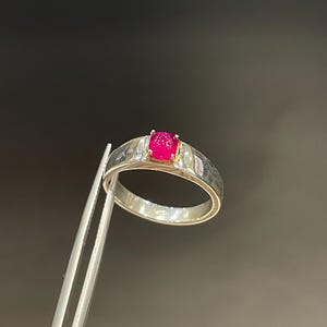 Könnte beinhalten: Ein silberner Ring mit einem rechteckigen, strukturierten Band und einem leuchtend roten Edelstein. Der ovale Edelstein ist in einer goldfarbenen Fassung gefasst. Der Ring wird von einer silbernen Pinzette vor dunklem Hintergrund gehalten.