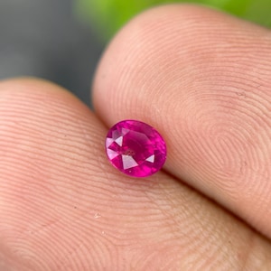 May include: A faceted, oval-shaped ruby gemstone. The gem is a vibrant shade of pinkish-red, with multiple facets catching the light. The ruby is held between two fingers, with a blurred green background.
